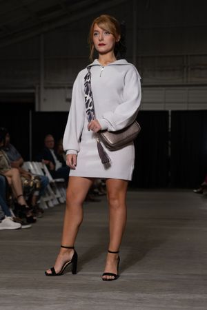 Model presents white hoodie dress with brown crossbody bag and black sandals on fashion runway.