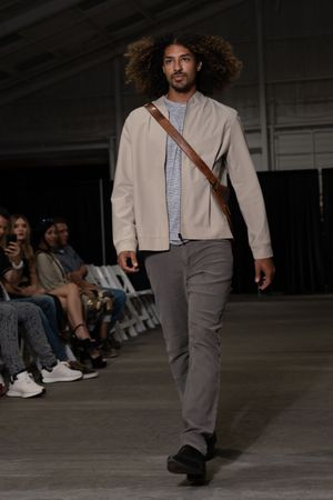 Model wearing white cropped jacket and gray pants walks down concrete runway during fashion show.