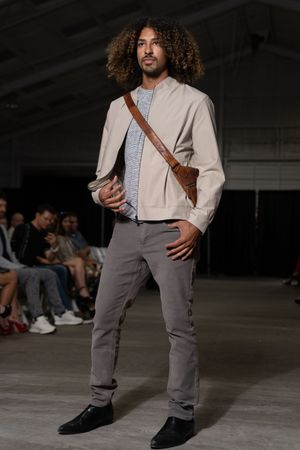 Model wearing beige jacket with brown messenger bag and gray pants on industrial catwalk.