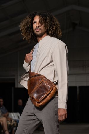 Model walks runway wearing beige blazer and grey pants while carrying brown leather handbag at fashion show.