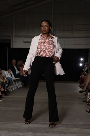 Model presents white blazer with patterned blouse and black flared pants on fashion runway.