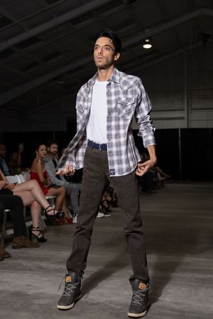 Model showcases casual streetwear with plaid shirt and dark pants on concrete runway.