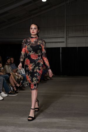 Model showcases fitted floral dress with long sleeves and black sandals on dark runway.