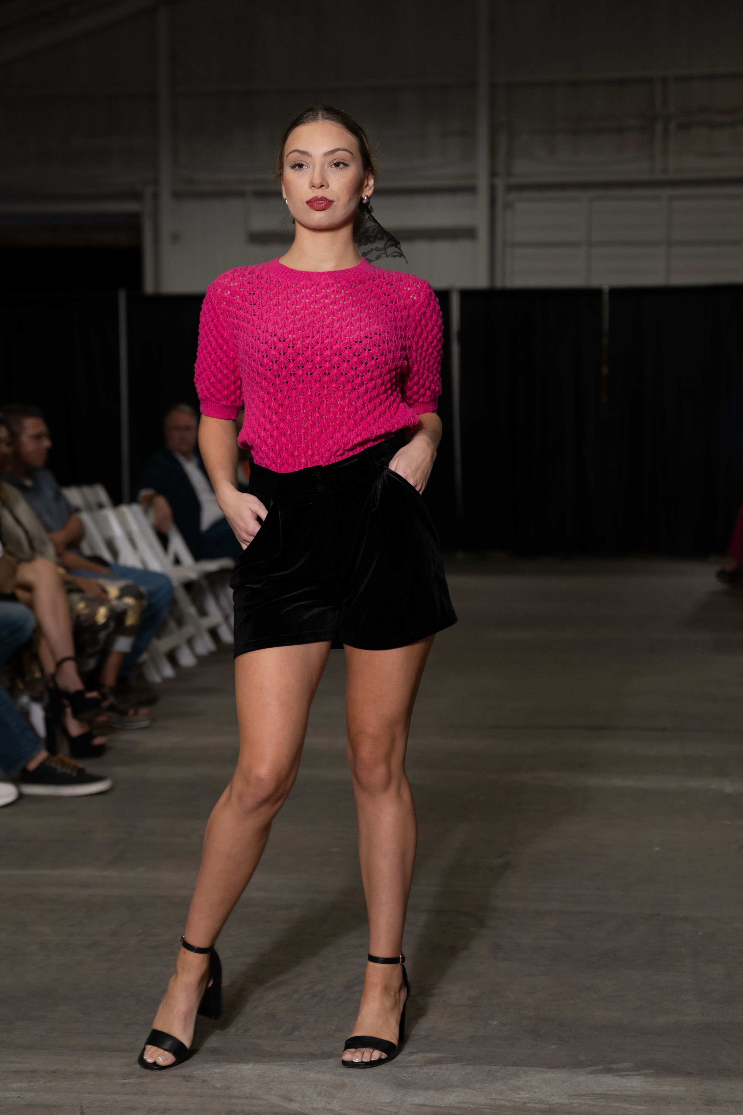 Model walks runway in bright pink asymmetrical top and black shorts during fashion show presentation.