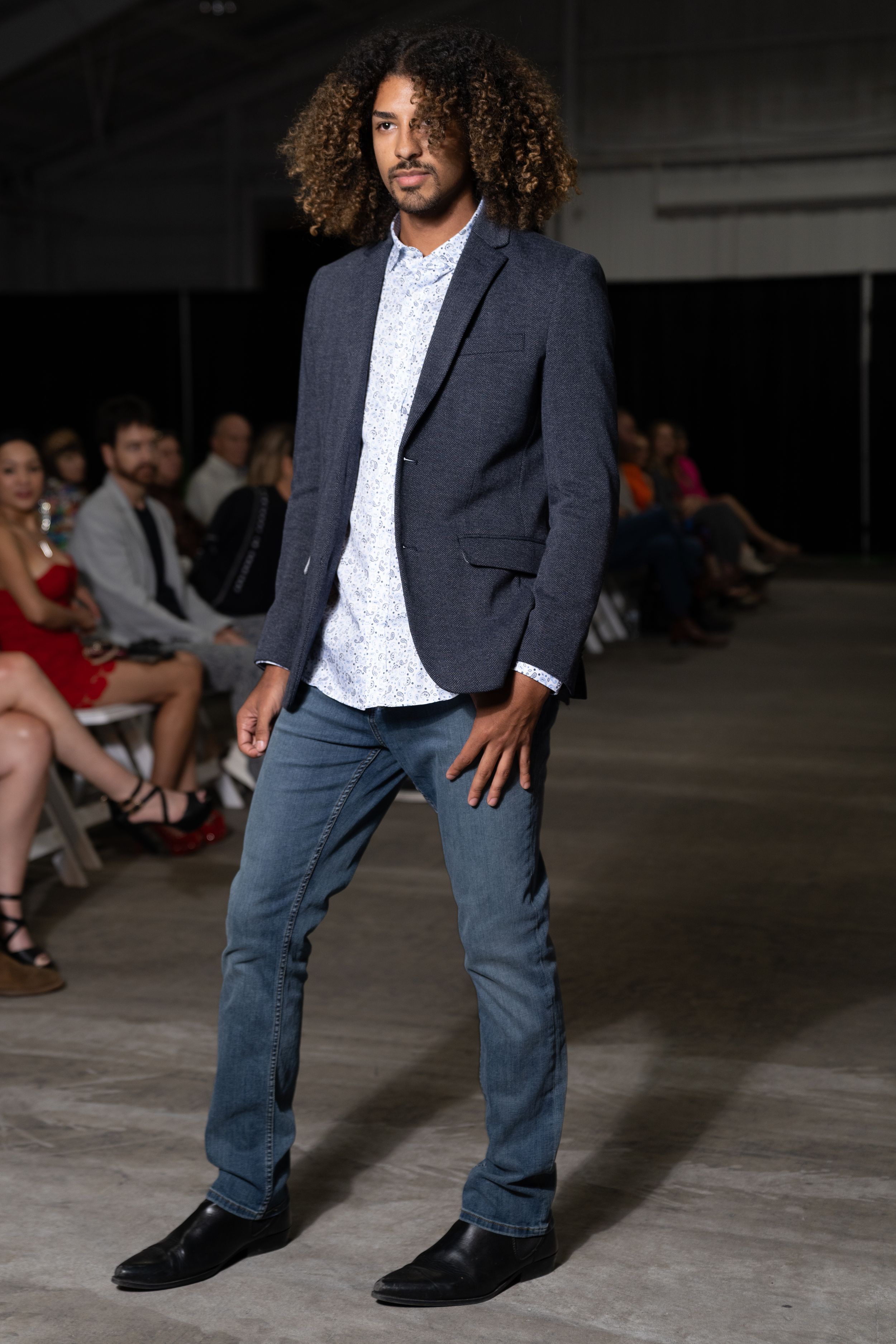 Model walks runway wearing navy blazer, white shirt and jeans during fashion show in industrial space.