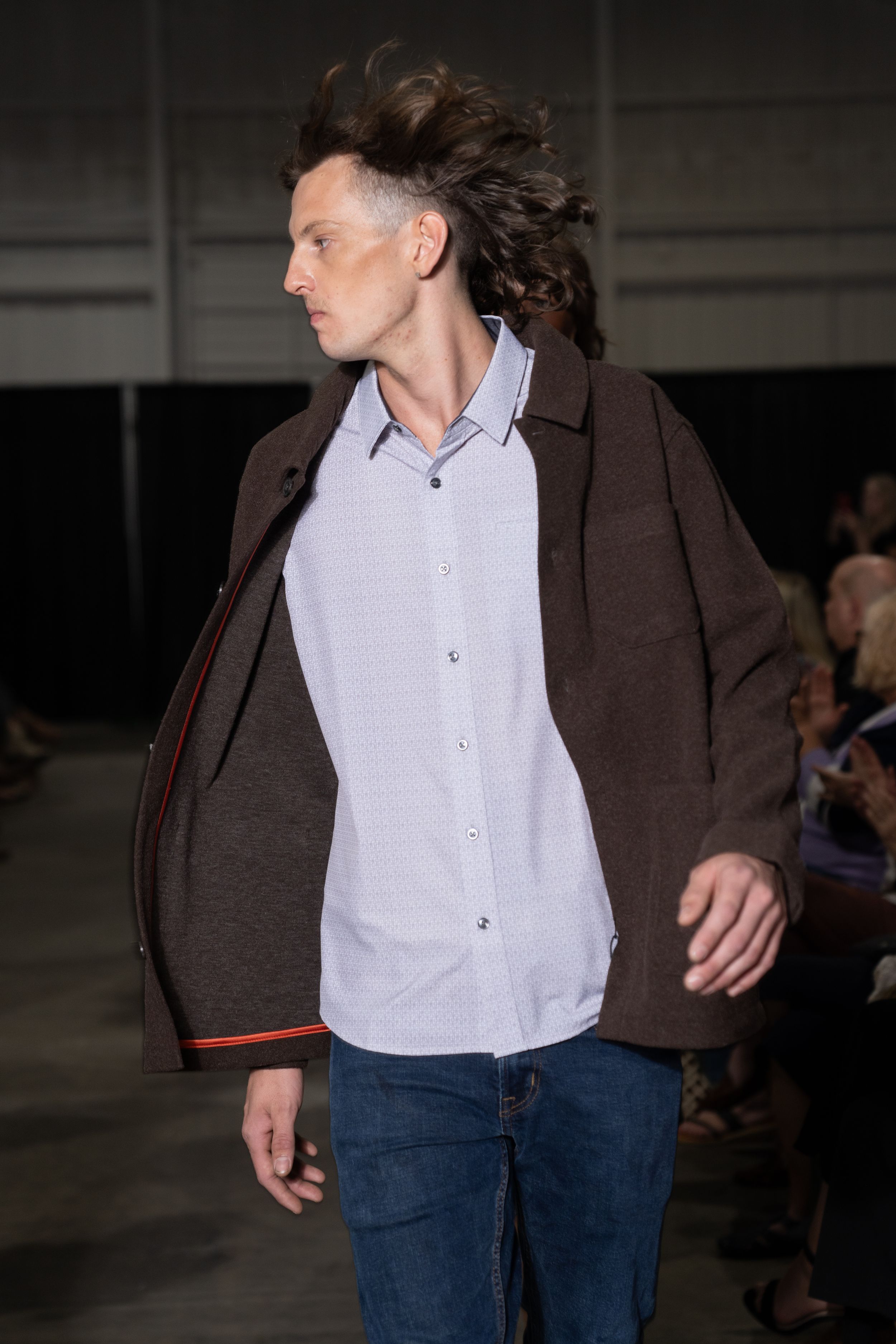 Model walks runway in casual outfit with brown jacket, white button-down shirt and jeans during fashion show.