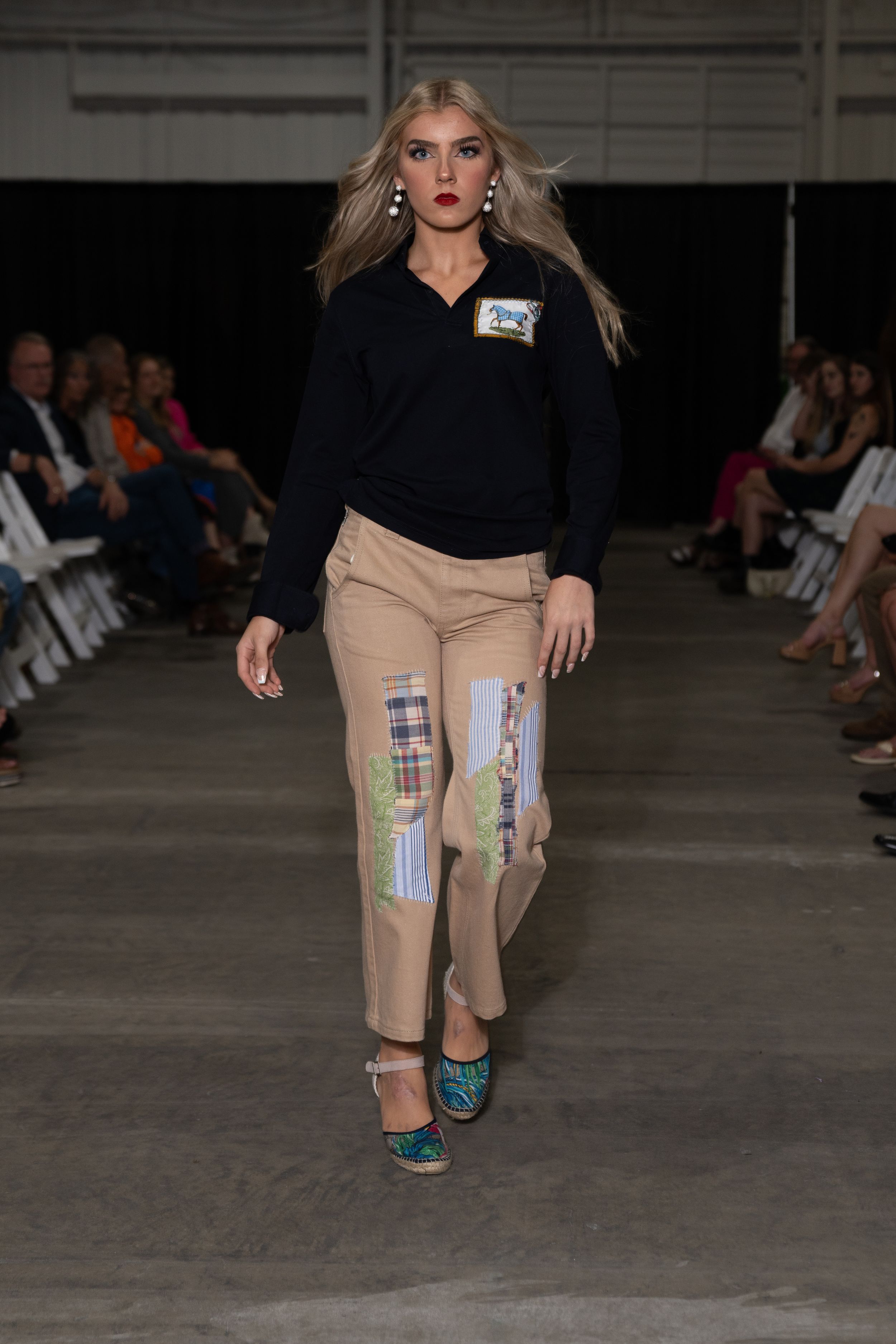 Fashion model walks runway wearing black top with khaki shorts and blue heels at underground fashion show.