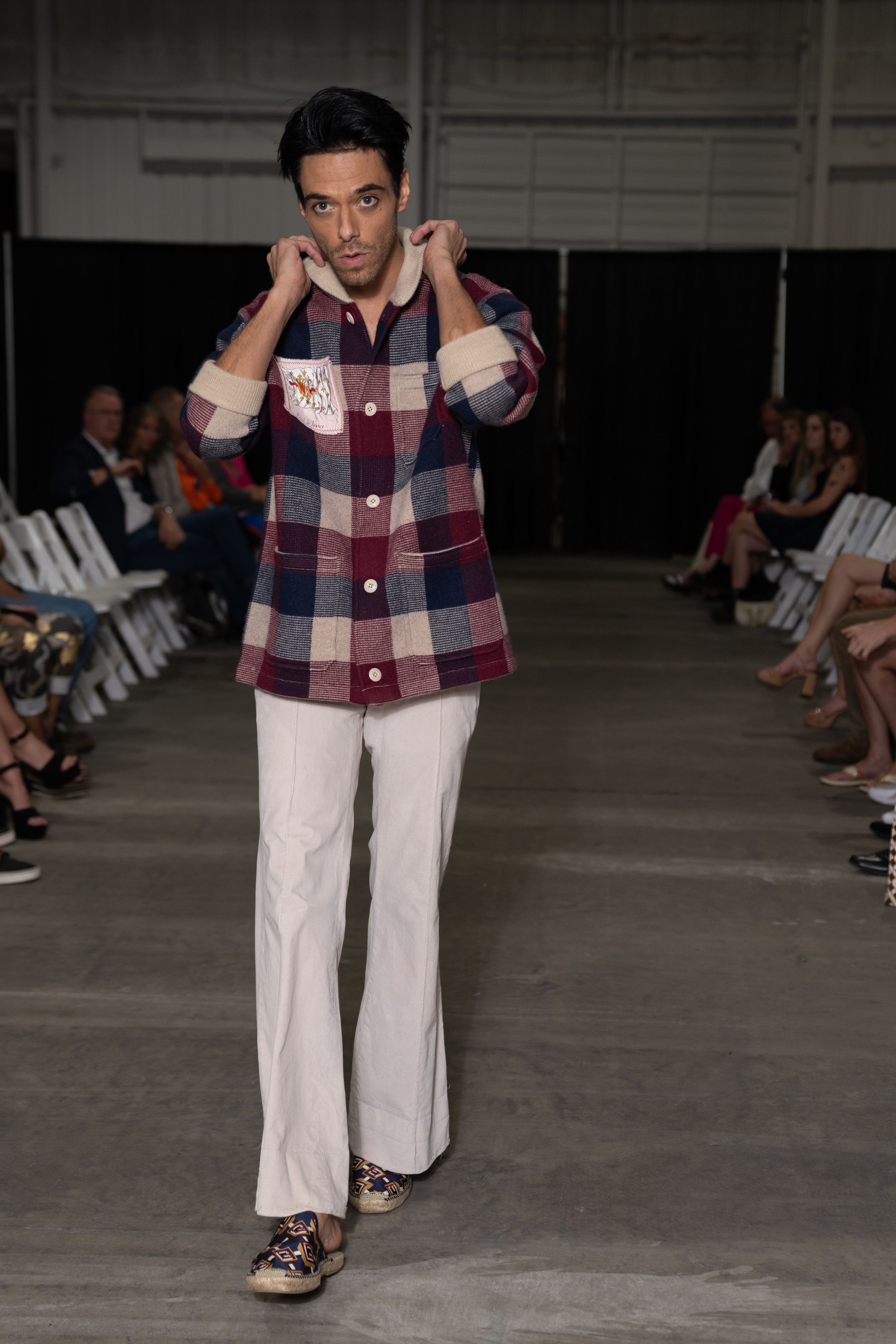Model showcases plaid jacket and white pants ensemble on concrete runway during fashion presentation.