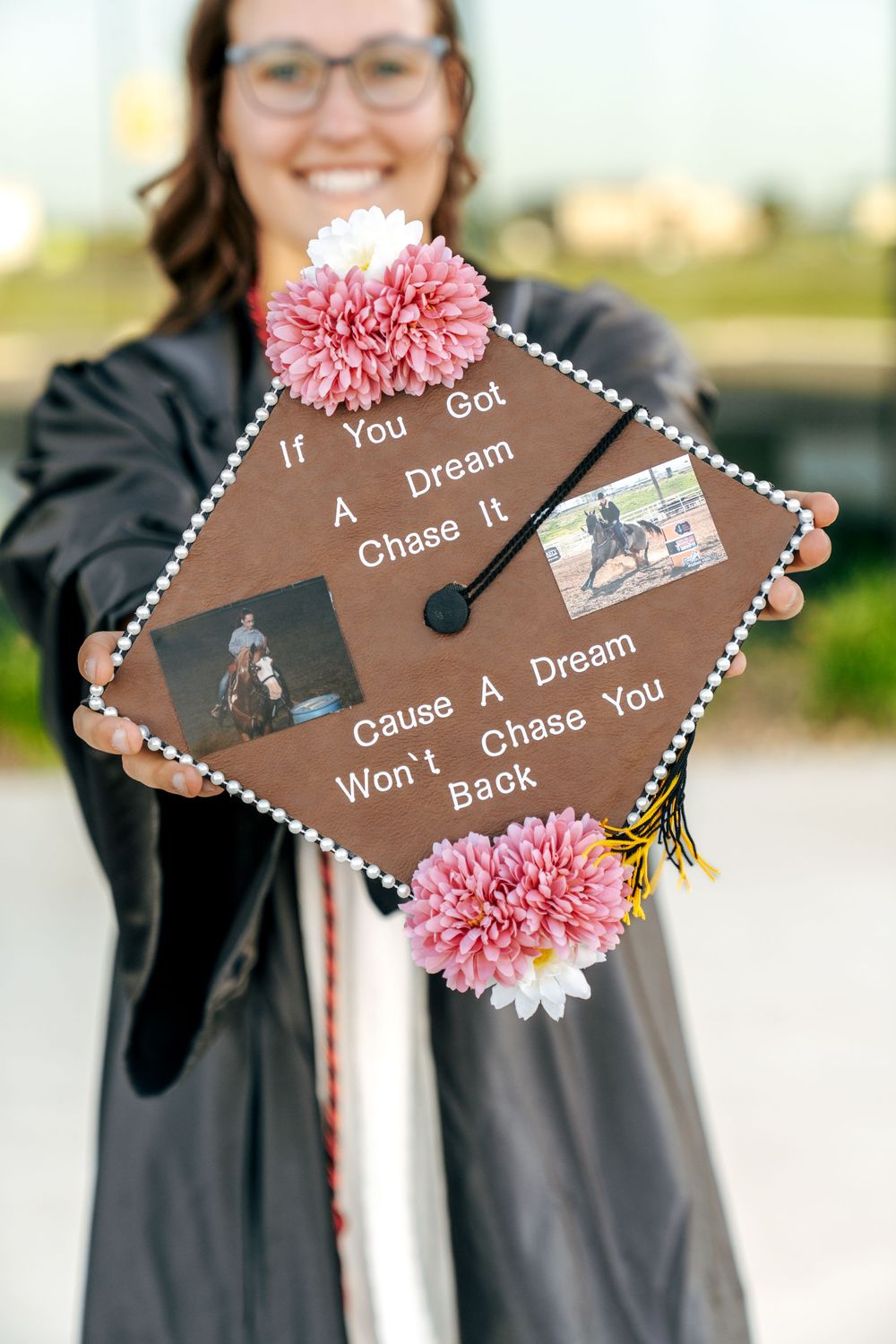 Graduate smiling in black gown holding decorated mortarboard with pink flowers and inspirational quote about chasing dreams.