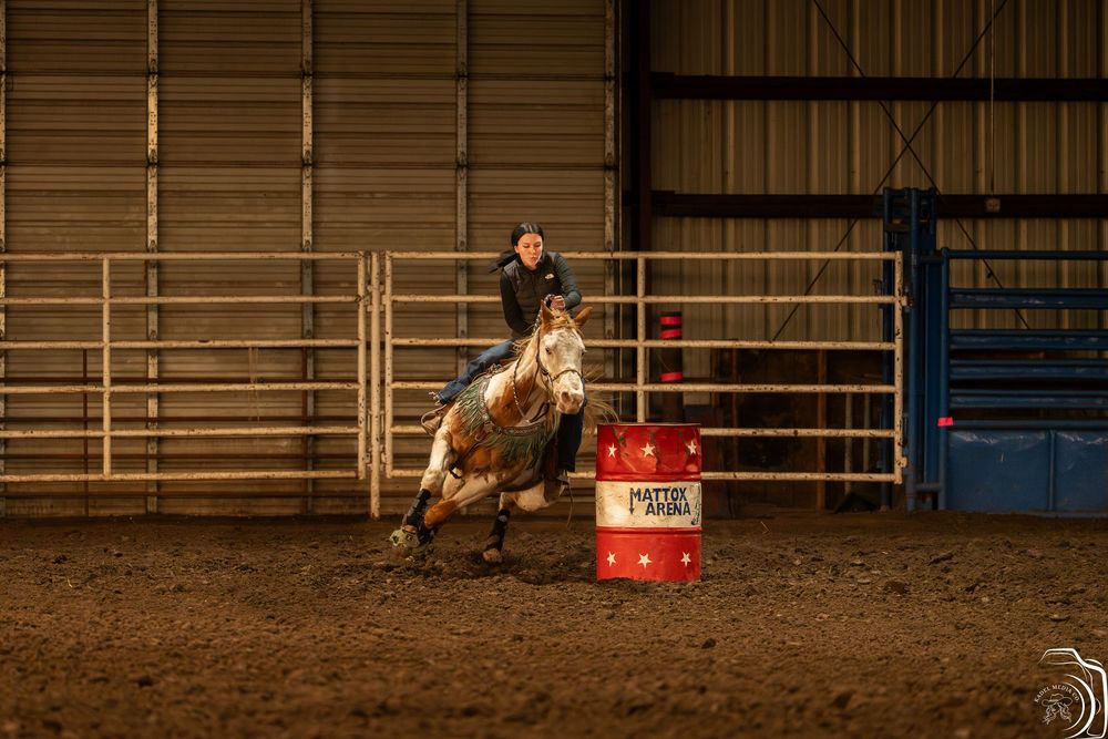 Barrel racer on a gray horse navigating a red barrel in an indoor rodeo arena during a competition.