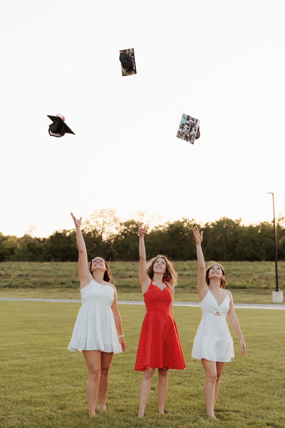 Three graduates in white and red dresses toss their graduation caps into the air outdoors.