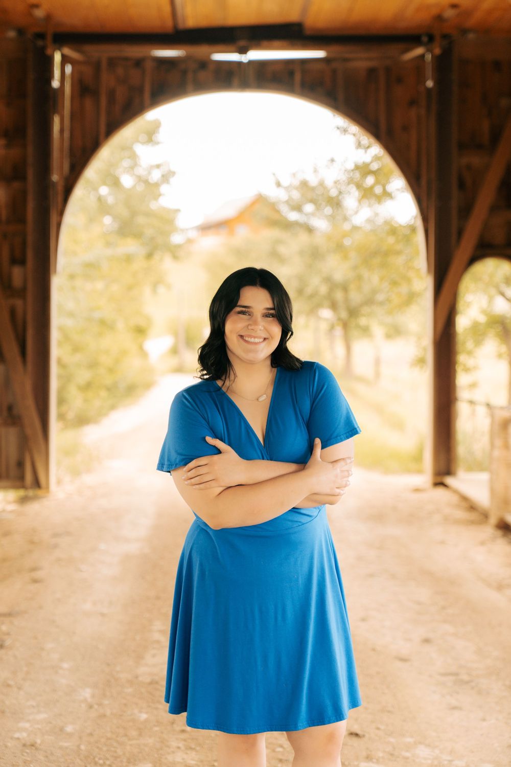 Smiling young woman in blue dress standing with arms crossed under a wooden arch bridge outdoors.