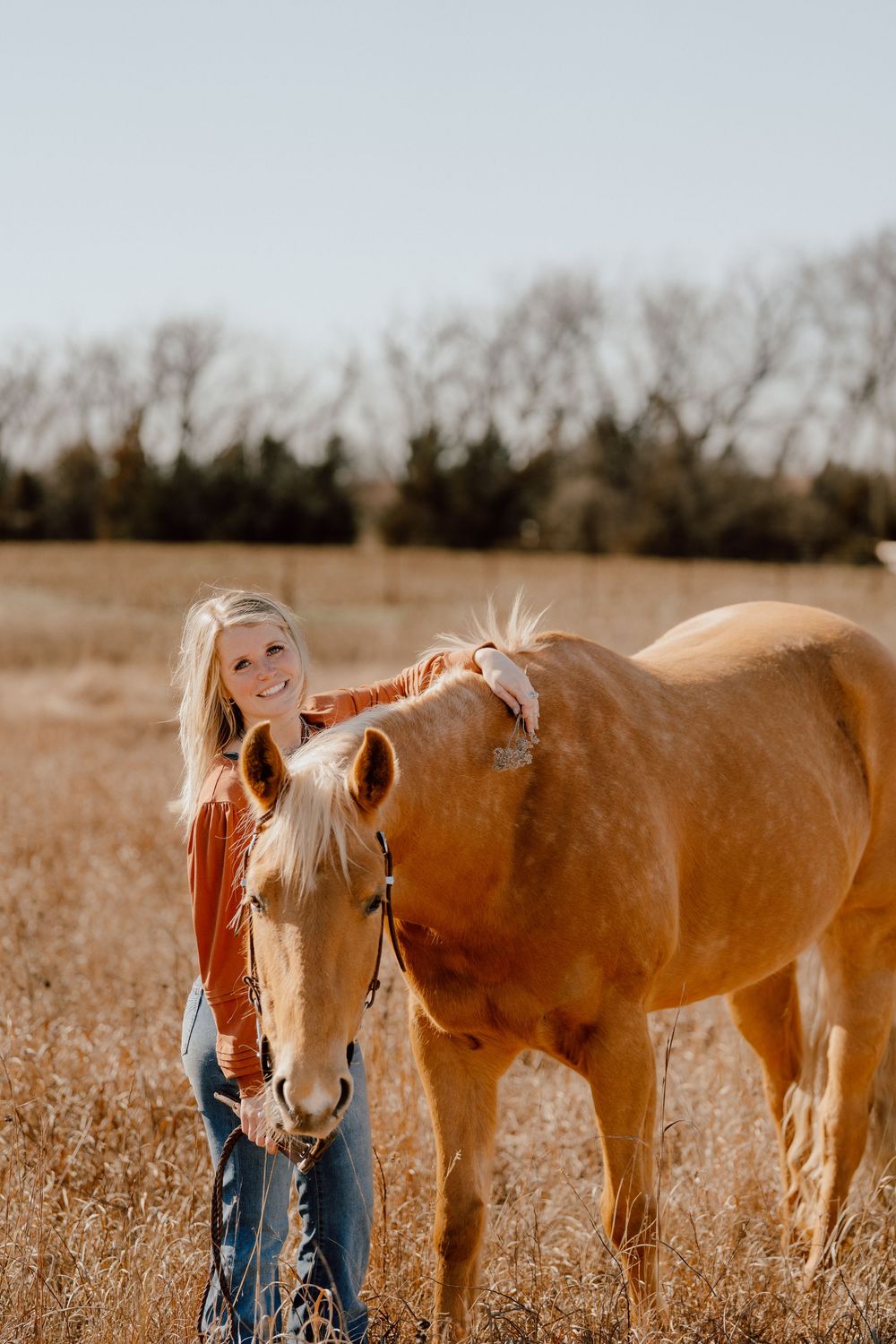 A smiling blonde woman in an orange top pets a golden palomino horse in a sunlit autumn field.