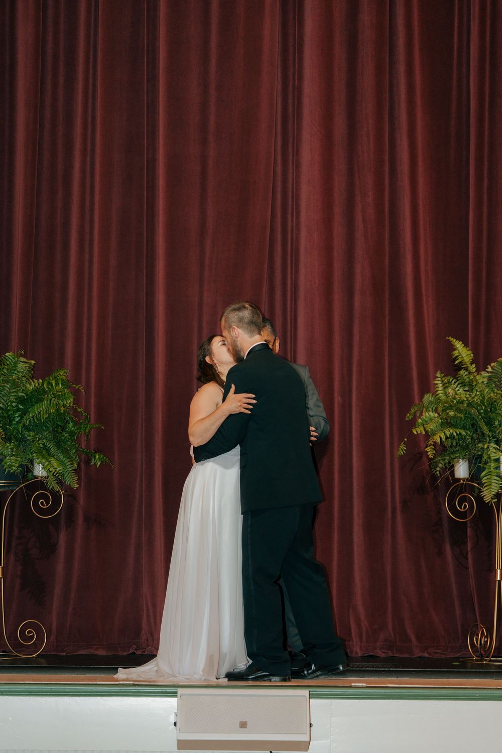 Couple sharing first dance kiss on stage with elegant burgundy velvet curtain backdrop and fern plants at wedding reception.