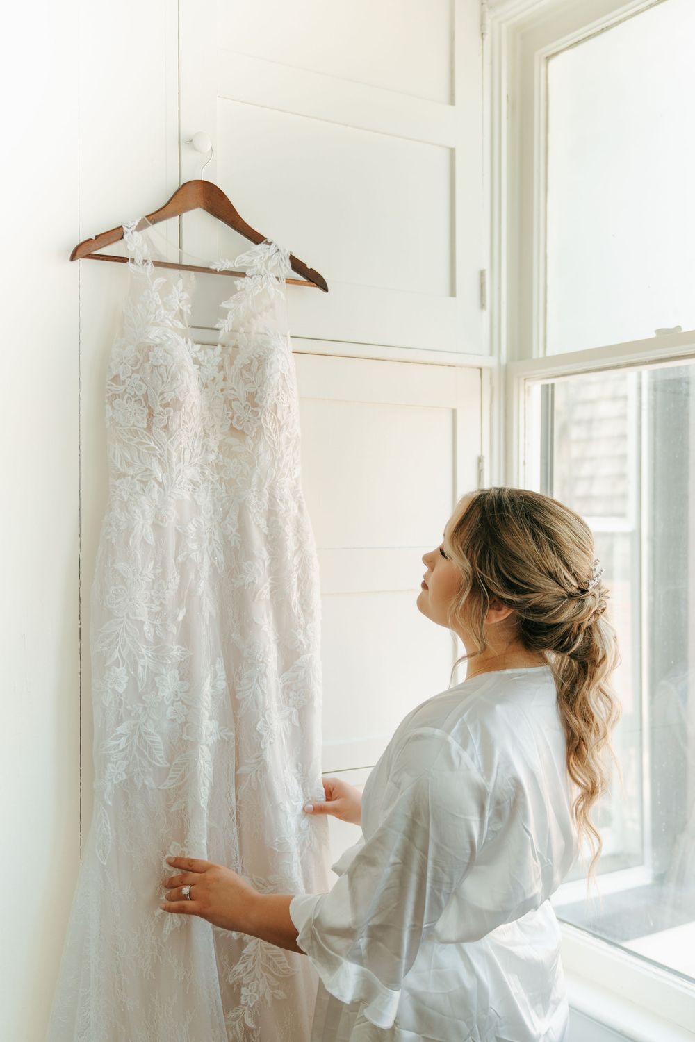 Bride in white robe admiring her lace wedding gown hanging by a bright window.