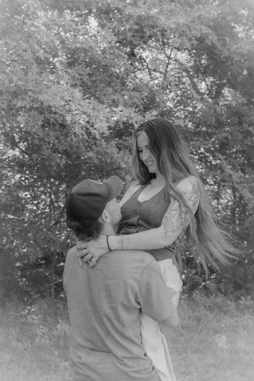 Black and white photo of a couple embracing outdoors, woman with long hair and tattoos smiling as man lifts her.