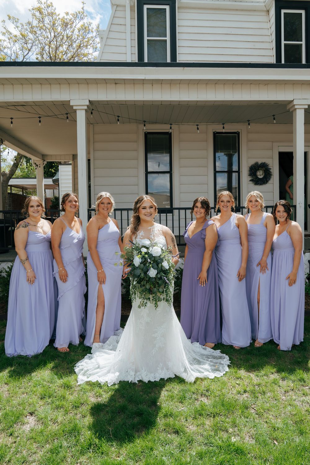 Bride with bridesmaids in lavender dresses posing in front of a white farmhouse venue.
