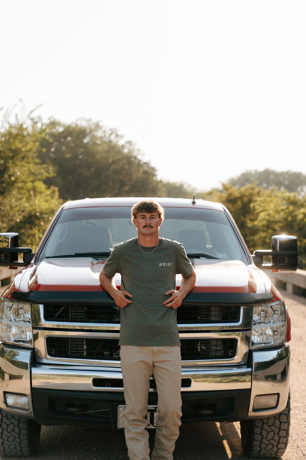 Young man with mustache in green Ariat tee and khakis leaning against a red and black Chevy pickup truck outdoors.
