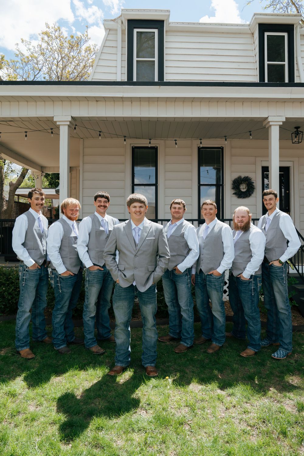 Groom and groomsmen in gray vests and jeans posing in front of a white farmhouse.