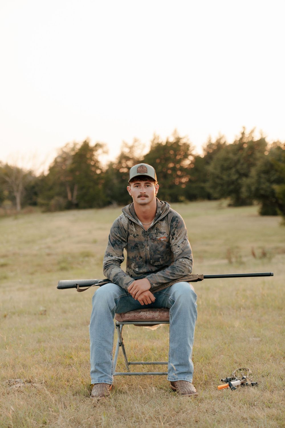 Young man in camo hoodie and cap sits in field chair holding rifle, with trees in background at dusk.