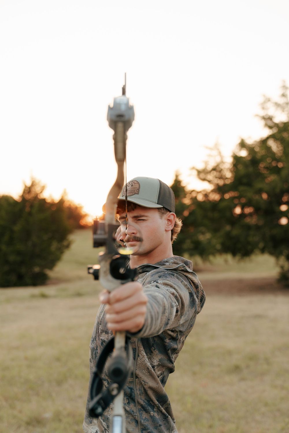 A young man in camouflage and a cap aims a compound bow outdoors at sunset.