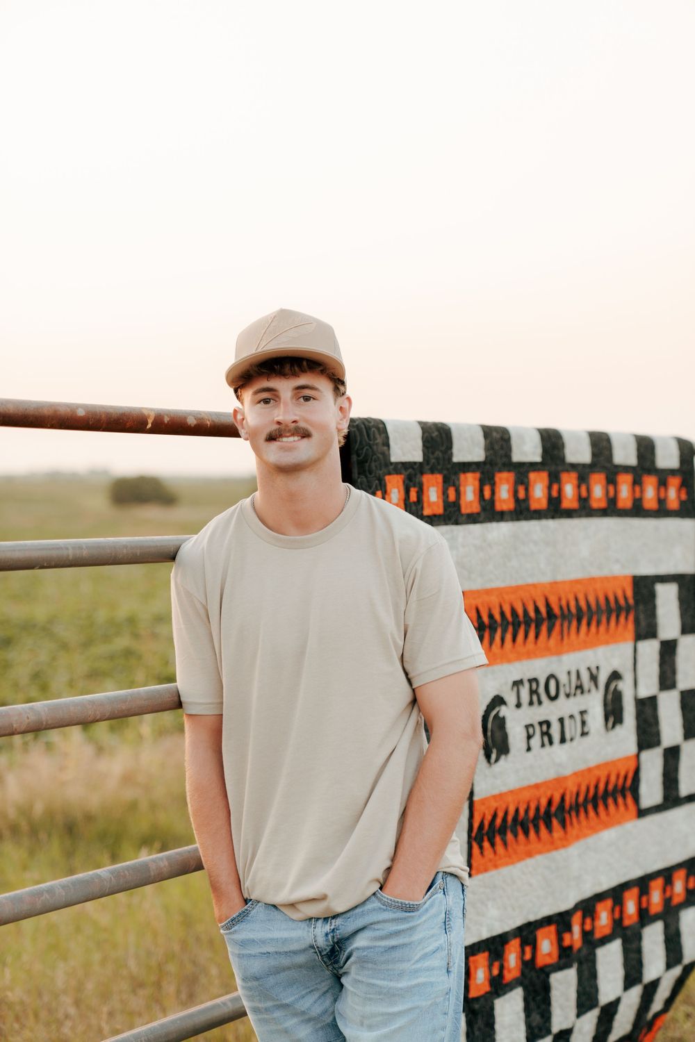 Young man with mustache in beige shirt and cap leaning on metal fence with Trojan Pride blanket outdoors.