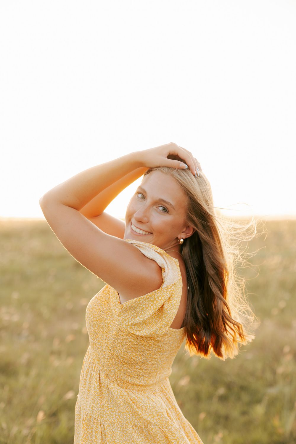 Young woman in yellow floral dress smiling in golden sunset field, hair blowing in wind.