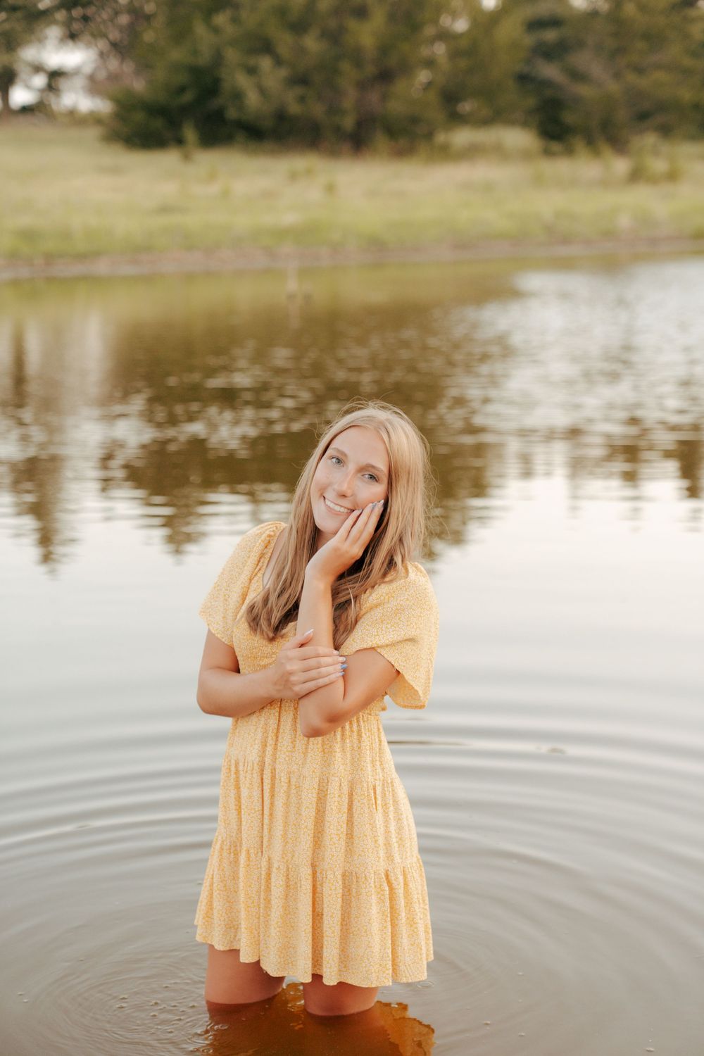 Young blonde girl in yellow dress standing in calm lake water, smiling with hand on cheek, trees reflected behind her.