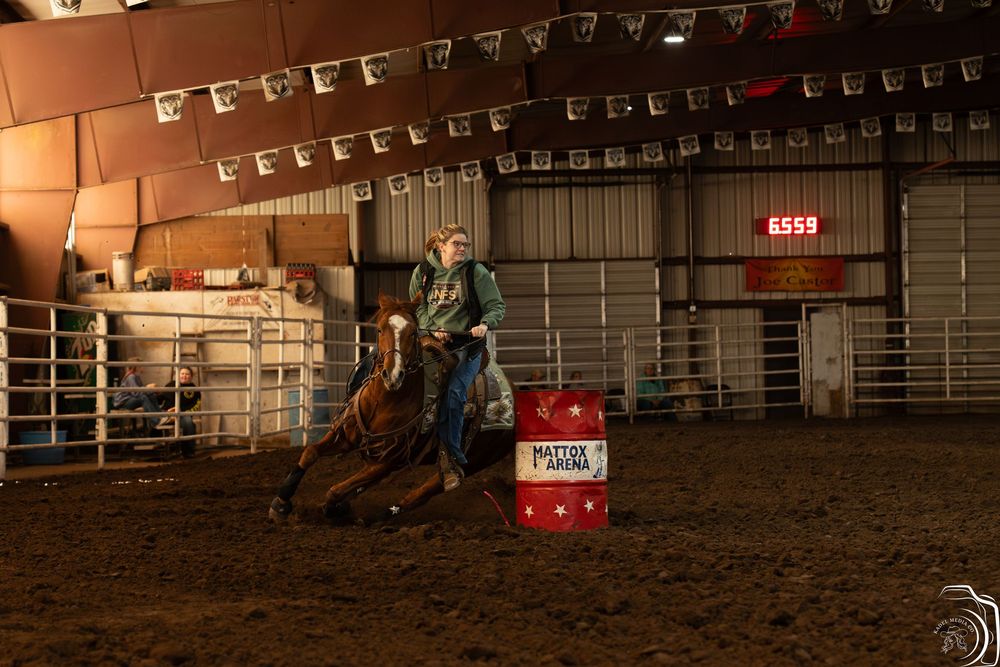 Barrel racer on horseback navigating tight turn around red barrel in indoor rodeo arena.