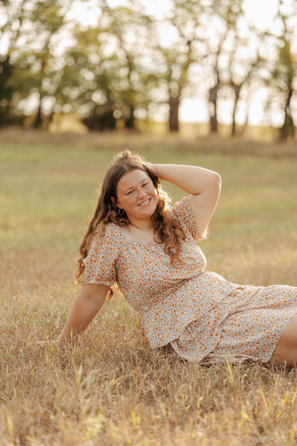 Young woman in floral dress sitting in golden field, smiling with hand in hair at sunset.