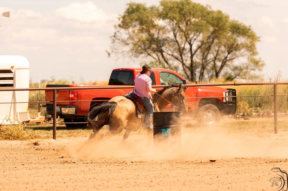 Rider on horseback performing barrel racing near red pickup truck on dusty ranch arena.