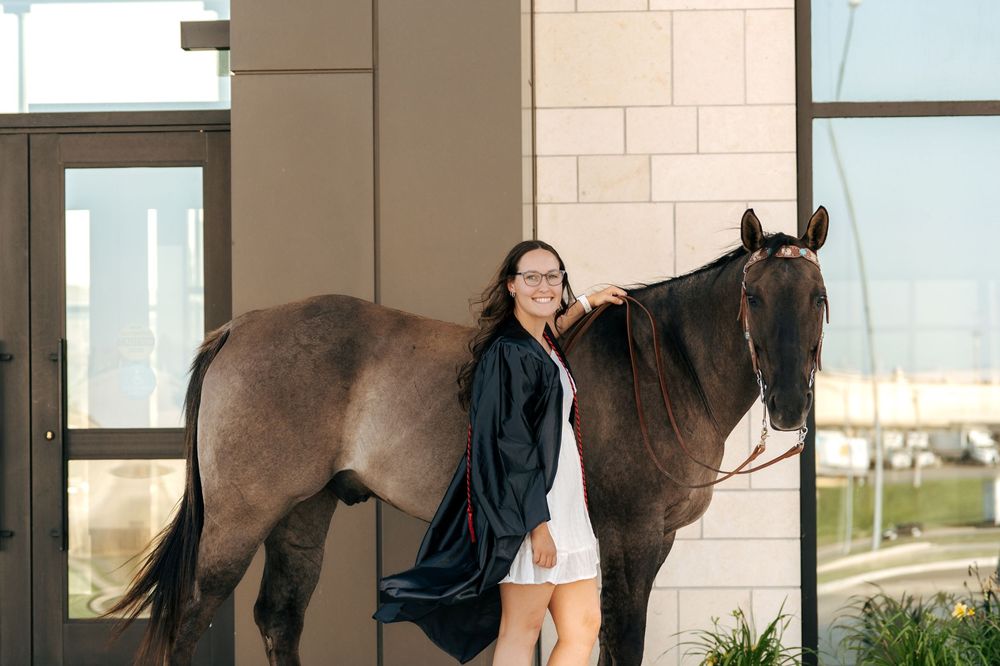 A smiling woman in graduation attire stands beside a dark brown horse outside a modern building.