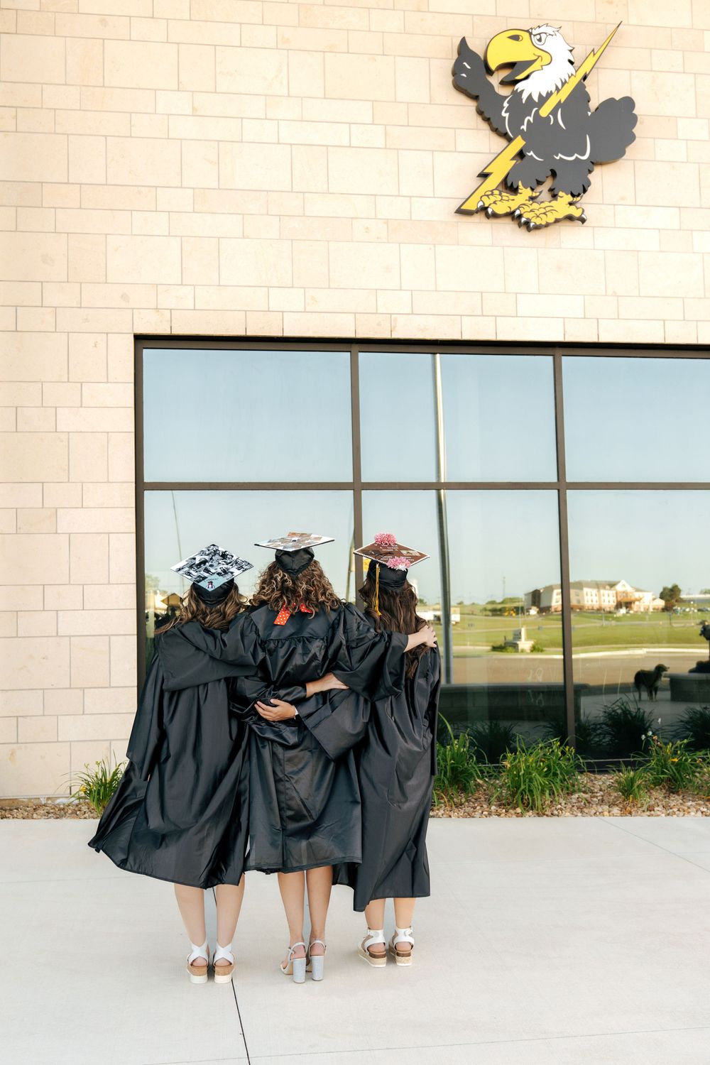 Three graduates in black gowns and caps stand together facing their school building with an eagle mascot logo.