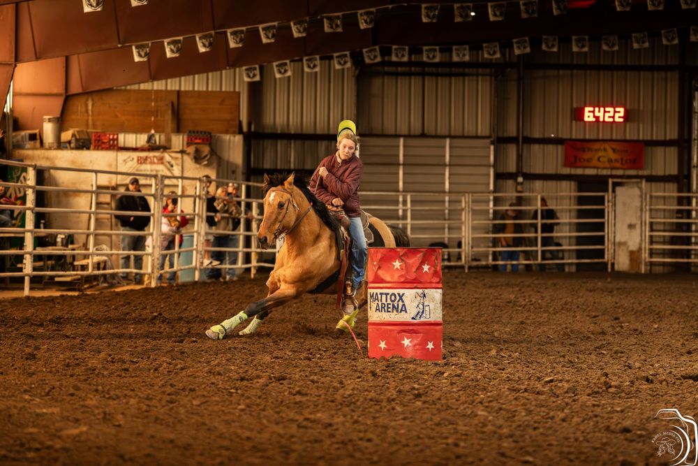 Young rider on a palomino horse competing in indoor barrel racing event at a rodeo arena.