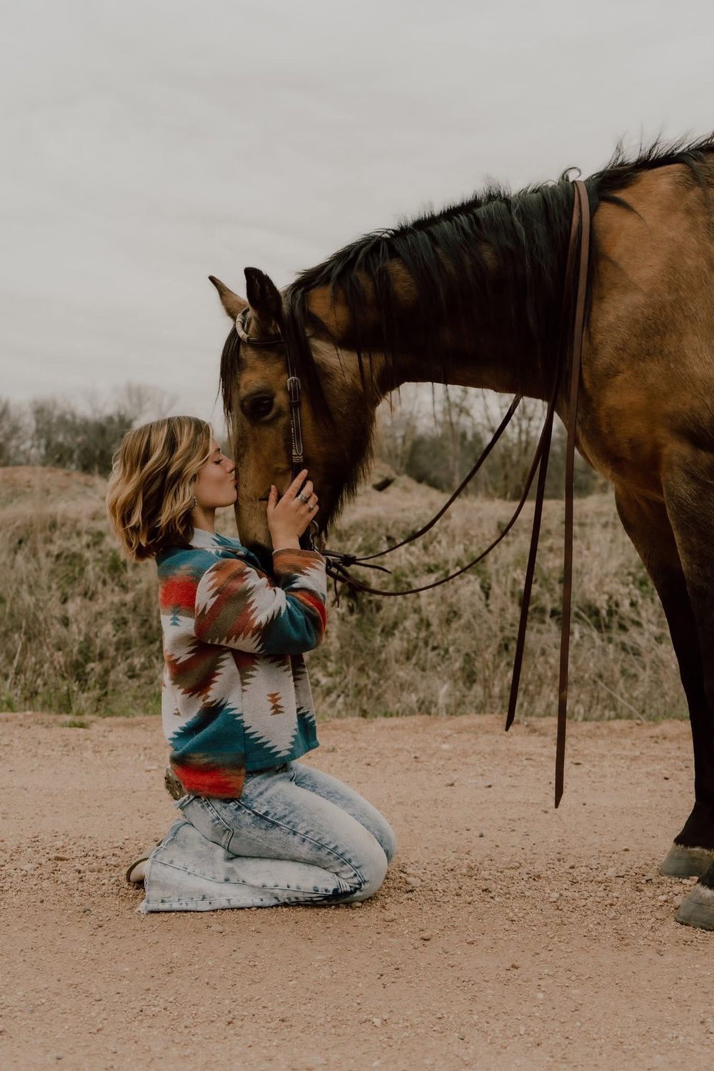 Young woman kneeling on the ground and affectionately nuzzling a brown horse outdoors.