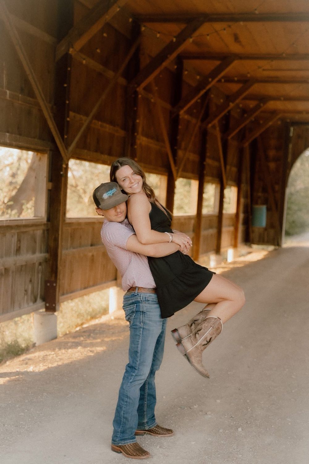 Couple embracing inside a rustic wooden barn, man lifting woman in a black dress and cowboy boots.