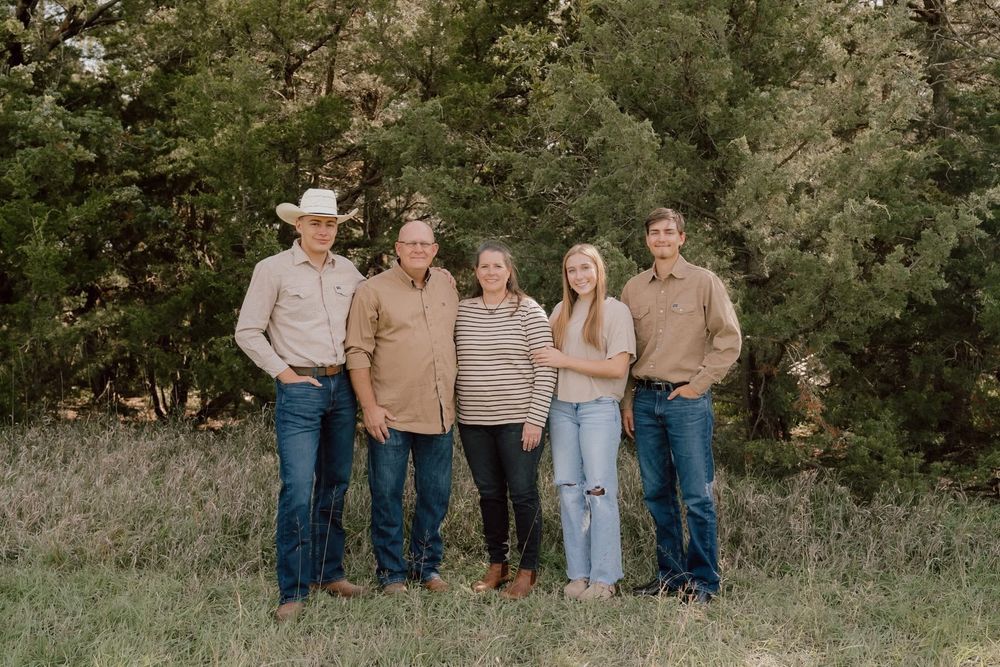 Family of five posing together in a field with cedar trees wearing coordinated tan and denim outfits.