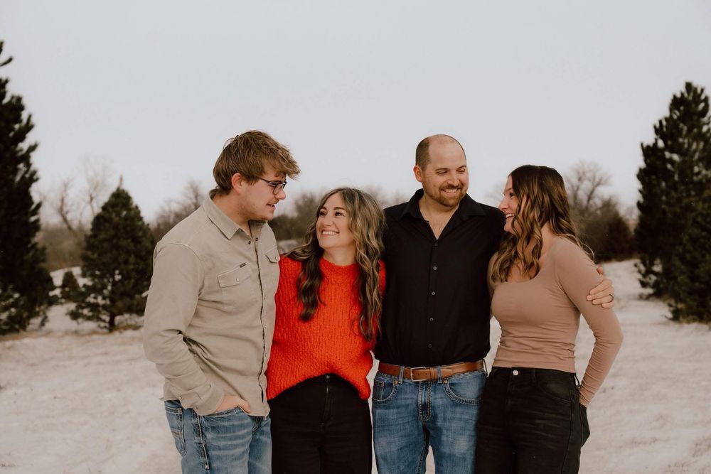 Group of four adults laughing and posing together in a snowy Christmas tree farm setting.