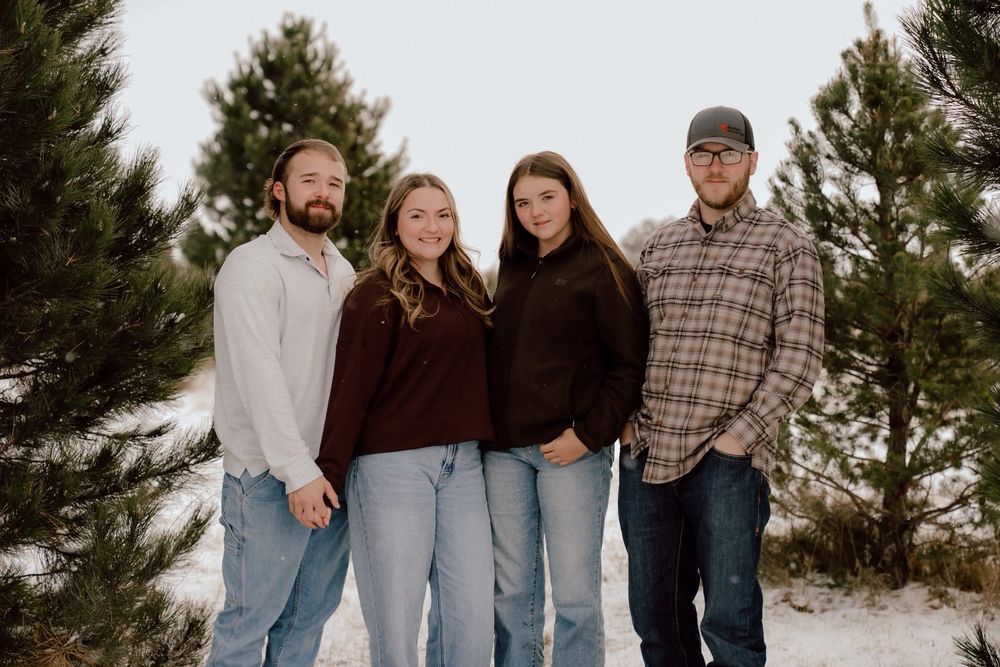 Four adults smiling together outdoors in a snowy pine tree setting wearing casual winter clothing.