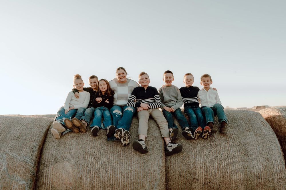 Group of nine children sitting together on large round hay bales under a clear sky.