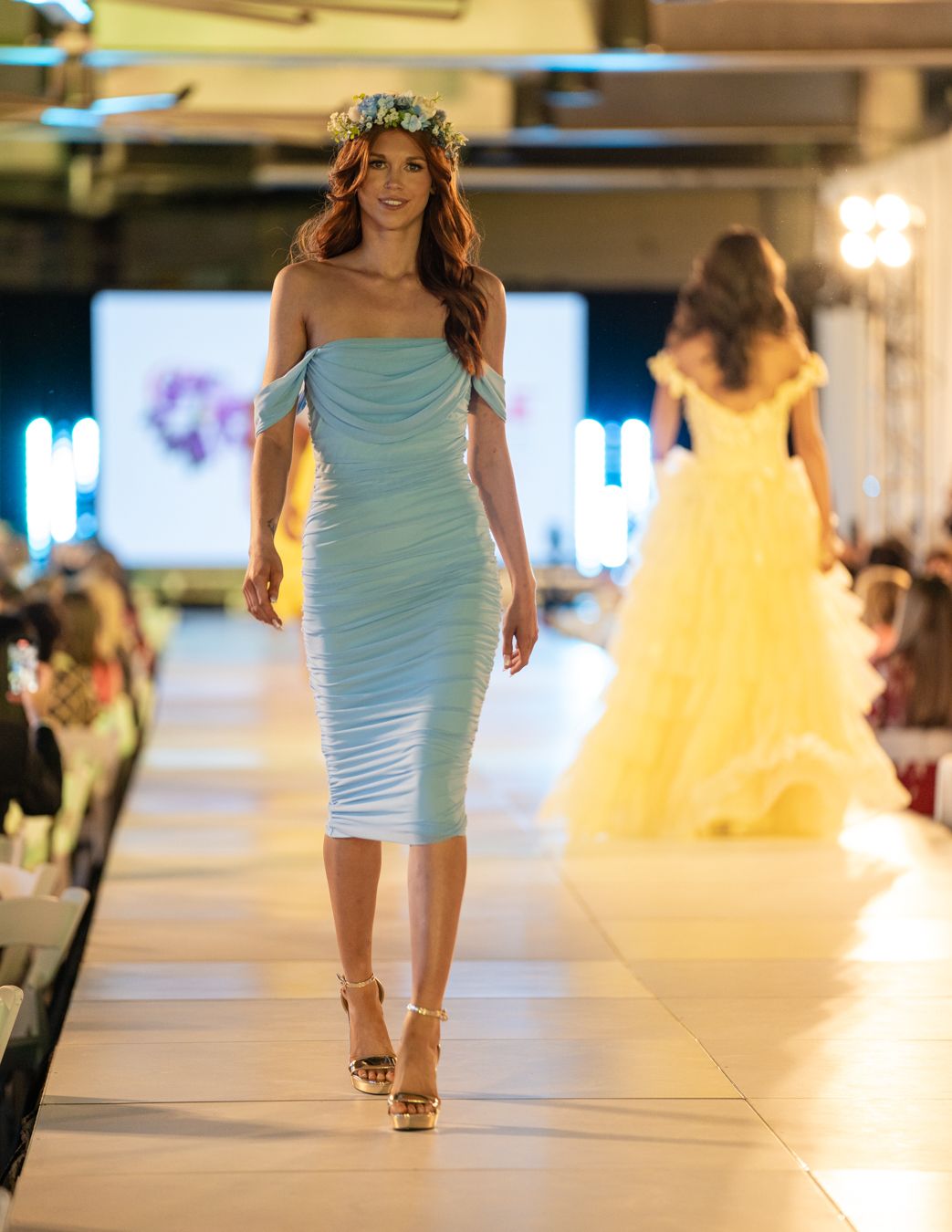 Model walks runway in light blue ruched off-shoulder midi dress and floral crown at fashion show with yellow gown in background.