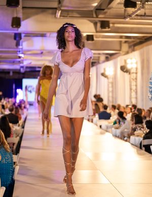 Models showcase summer fashion on a brightly lit indoor runway with white flooring and audience seating.