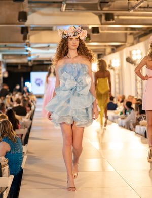 Models showcase light blue ruffled designs on a bright indoor runway with audience members seated along the sides.