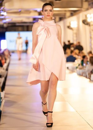 Models display light pink flowing garments while walking down a brightly lit indoor fashion runway.