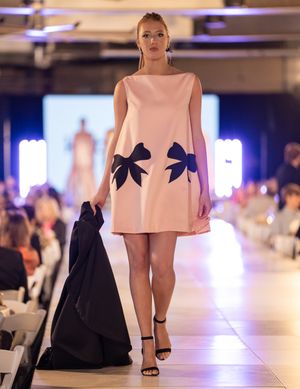 Model walks runway in pink mini dress with black bow details during fashion show.