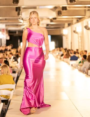 Models present vibrant pink evening gowns on a well-lit fashion runway with audience in background.