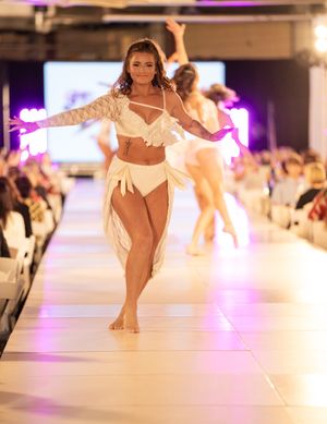 Fashion models displaying white swimwear collection on illuminated catwalk with purple lighting effects.