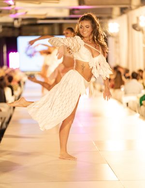 Models showcase flowing white dresses on an illuminated runway during a fashion show.
