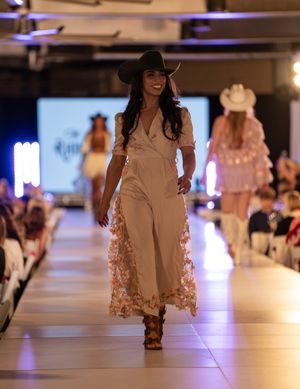 Model walks runway in beige western-inspired jumpsuit with floral details during fashion show.