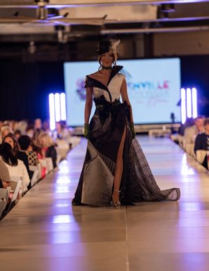 Models walk the runway in elegant black evening gowns at a fashion show with dramatic purple lighting.
